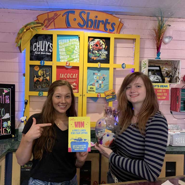 Two woman holding a ticket and a bowl for a raffle that an office can win to have a free lunch on Chuy's.